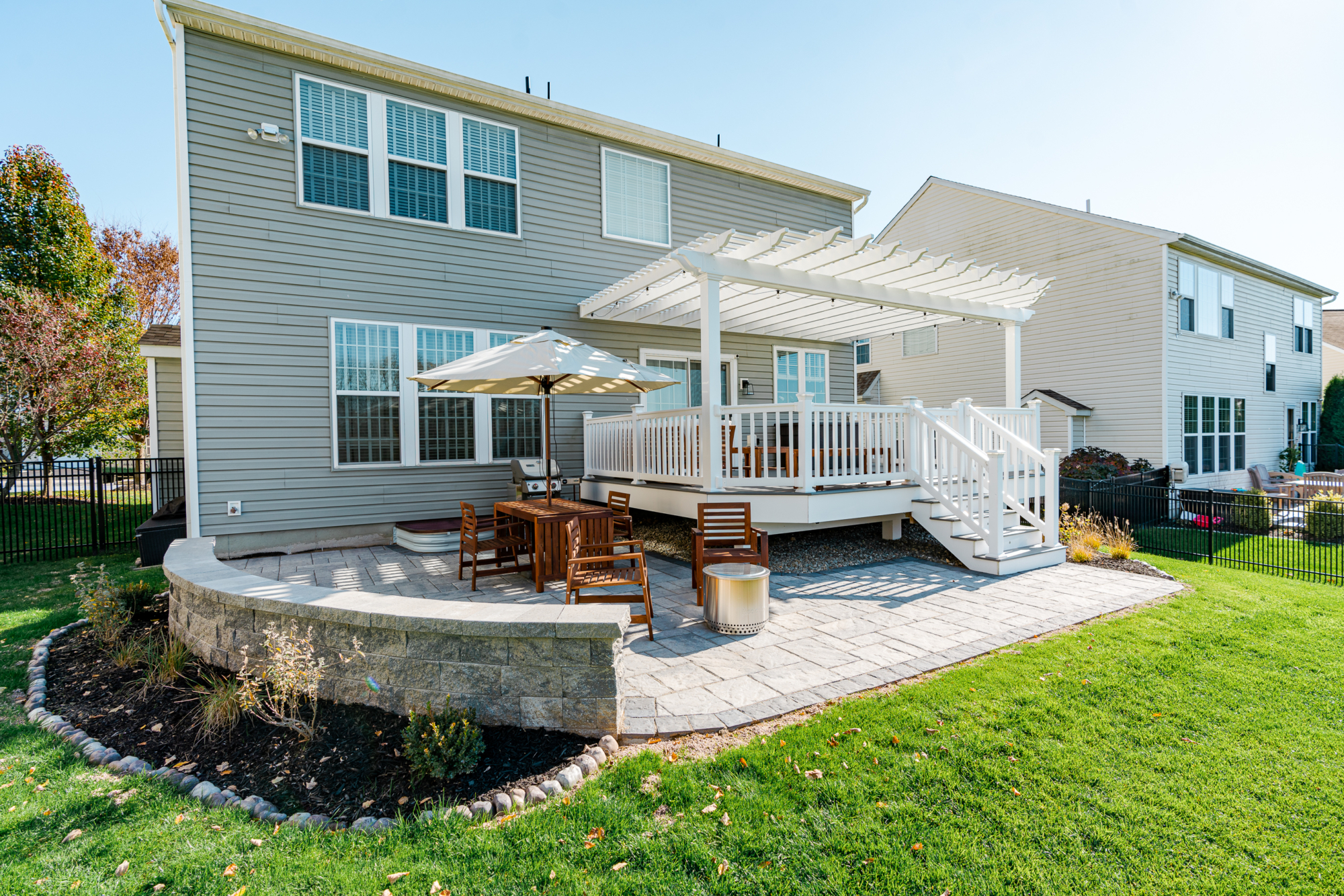 image of a deck and patio in downingtown pa built by integrous fences and decks featuring a white vinyl pergola timbertech decking white vinyl railing paver patio and sitting wall around the yard is a black aluminum fence