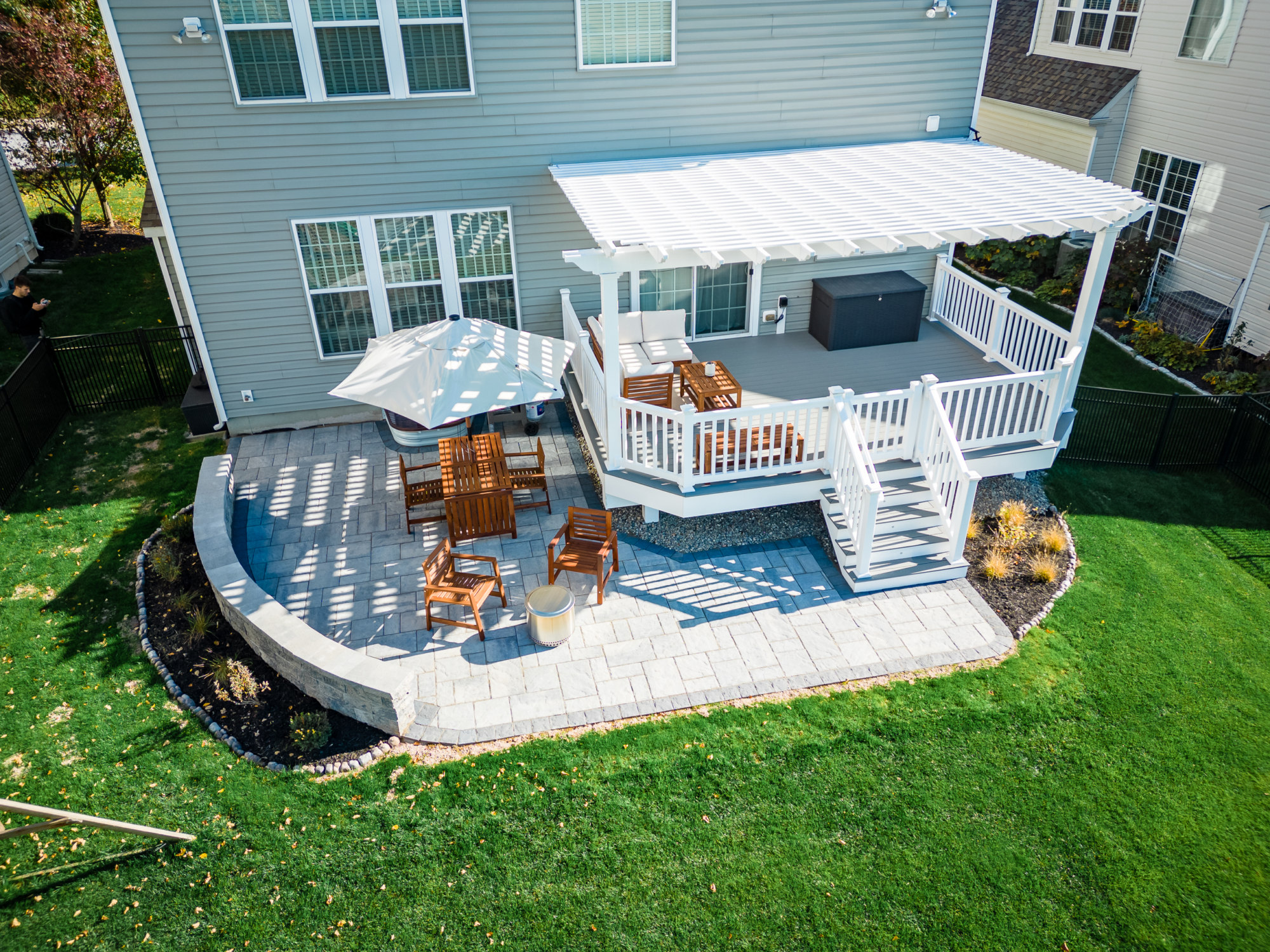 image of a deck and patio in downingtown pa built by integrous fences and decks featuring a white vinyl pergola timbertech decking white vinyl railing paver patio and sitting wall around the yard is a black aluminum fence